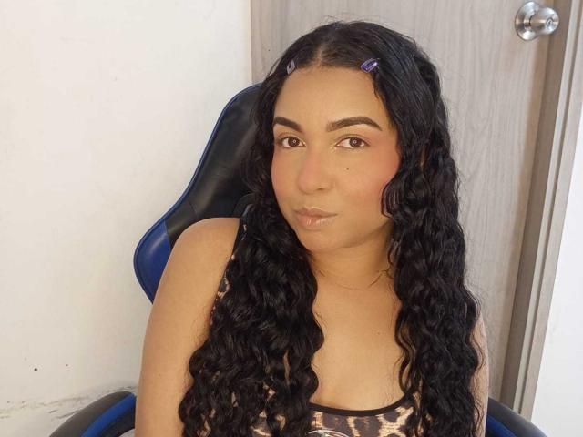 Photograph of AliceSmity with long, wavy black hair, sitting in a blue chair, pouting slightly, wearing a black and white patterned tank top, wooden door in background. Fucking sexy.