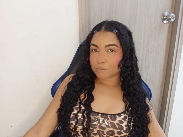 AliceSmity, a chubby woman with long, curly black hair, wearing a leopard-print tank top, sits in a blue chair, pouting with plump lips, and brown eyes. Silver door handle in background.