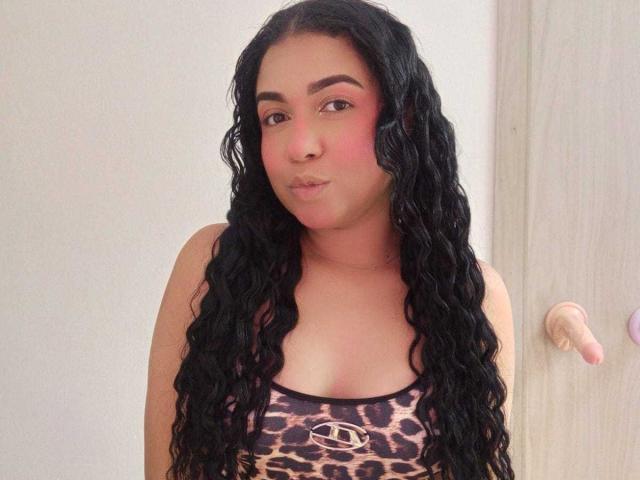 Fotografia de Alice Smity, uma mulher curvilínea com cabelos longos e ondulados, pretos, vestindo uma regata estampada com onças. Ela tem um blush rosa nas bochechas, e seus lábios franzidos sugerem algo. Seus olhos estão semicerrados..