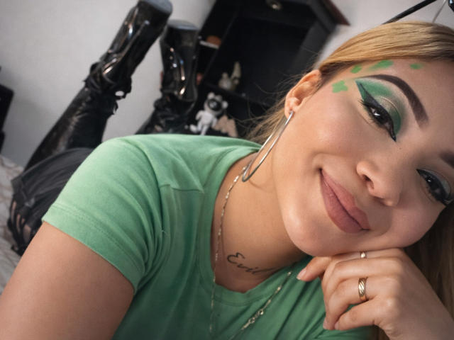 Primo piano di TatianaRossi con trucco verde per gli occhi, una camicia verde e piccoli punti di trucco verde sul viso. Lei sorride, appoggiando la testa sulla mano, mostrando un anello al dito. Incredibilmente attraente..