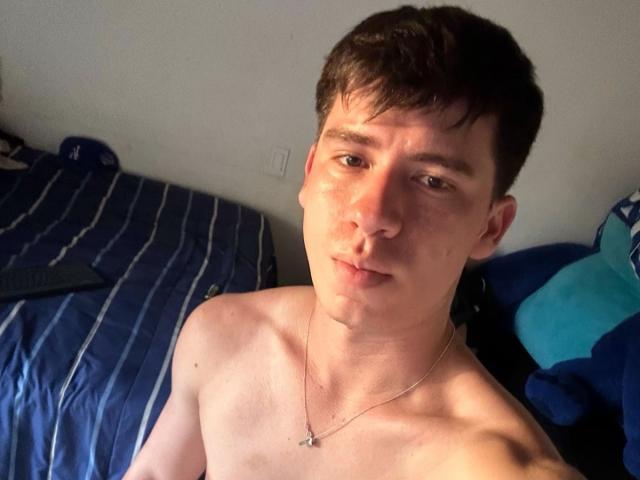 Fotografia de um jovem sem camisa, Sebastia Quesada, com cabelo curto e escuro, pele clara e um colar, sentado em uma cama com listras azuis, olhando para a câmera com uma expressão neutra. Sombras claras no peito.