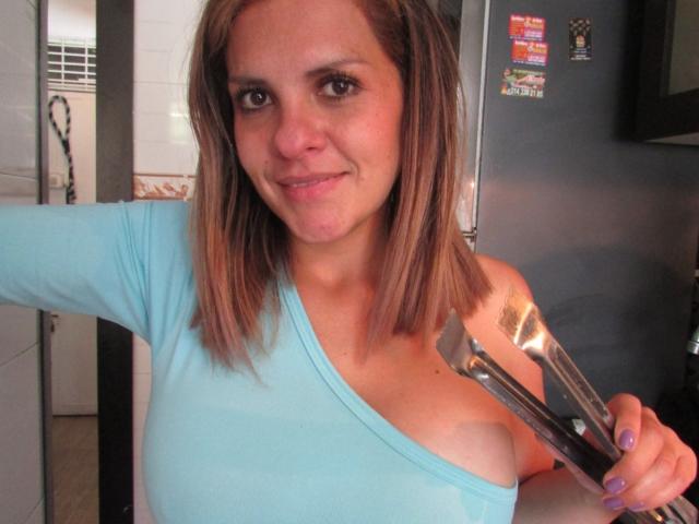 AbbyySmiith, une femme brune avec des cheveux mi-longs, portant un haut bleu clair à une seule épaule, tenant une pince de cuisine près d'elle. Sein droit, avec un léger sourire, réfrigérateur de cuisine avec des aimants en arrière-plan. Tension sexuelle évidente..