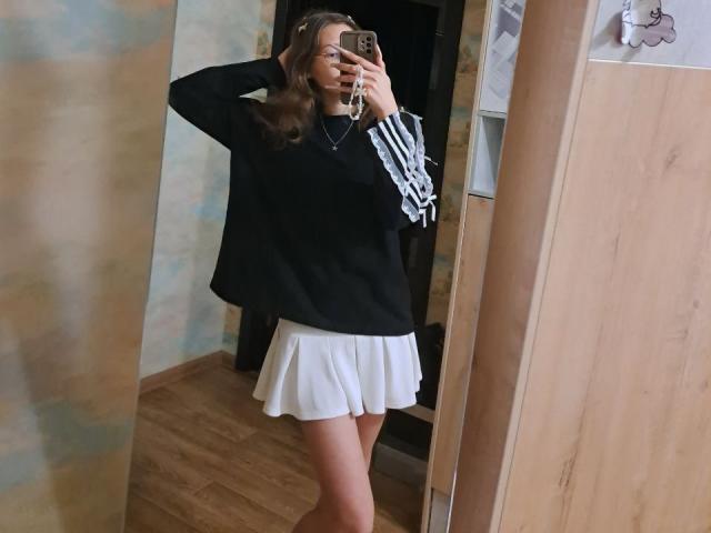 DelfinaAuble tira uma selfie no espelho, vestindo um suéter preto grande e uma saia branca curta plissada, segurando um celular. de um lado, cabelo castanho, chão de madeira, pose casual, leve tensão sexual..