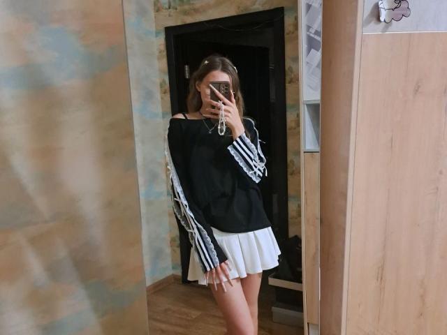 DelfinaAuble se toma un selfie frente al espejo, sosteniendo un teléfono con una mano. Ella lleva una camiseta negra de manga larga con rayas blancas y una falda blanca corta. Está de pie con una postura ligeramente provocativa..