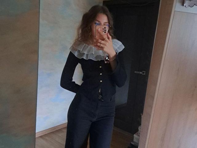 Delfina Auble, mit welligem, braunem Haar, trägt ein schwarzes Hemd mit Knopfleiste und einen weißen, gerafften Kragen und macht ein Selfie vor einem Spiegel. Sie legt eine Hand an ihre Hüfte und betont damit ihre weiblichen Rundungen..