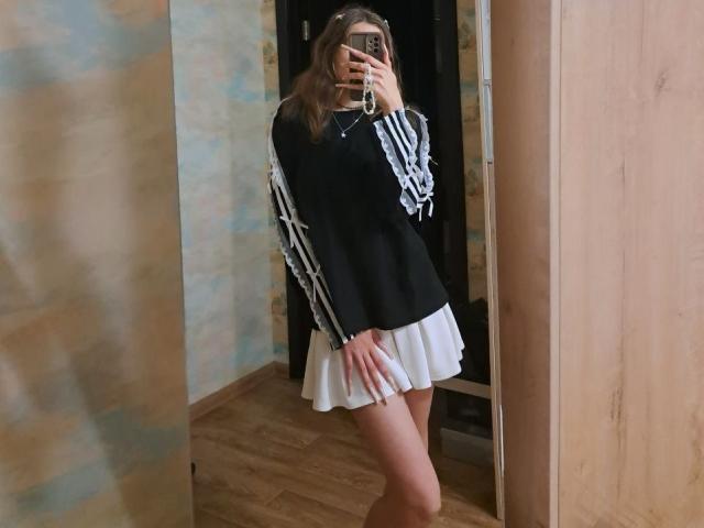 Delfina Auble è in piedi di fronte a uno specchio, coprendosi il viso con un telefono. Indossa una maglia nera a maniche lunghe con strisce bianche e una gonna bianca plissettata. Pavimento e pareti in legno. Incredibilmente sexy..