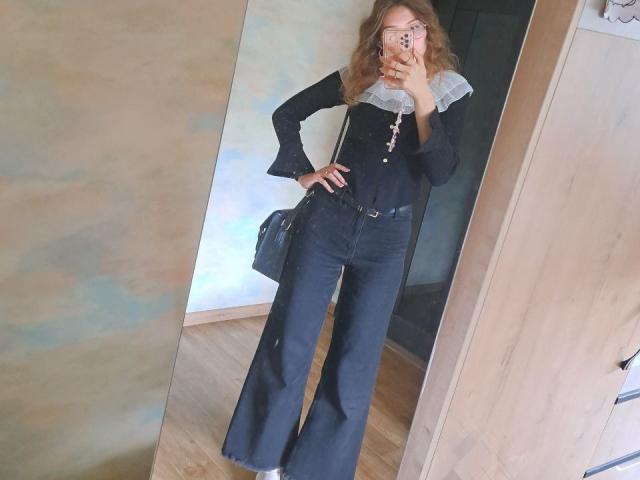 Delfina Auble è in piedi di fronte a uno specchio, indossa una camicetta nera con un bordo in pizzo bianco e dei jeans neri a vita alta. Lei tiene un telefono, che copre parte del suo viso. La sua mano è appoggiata sul fianco..