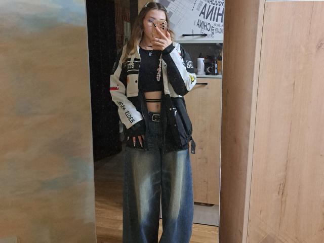 DelfinaAuble stands in front of a mirror, holding a phone to her face. She wears a black crop top with cutouts, loose blue jeans, and a black-and-white jacket. Fucking sexy.
