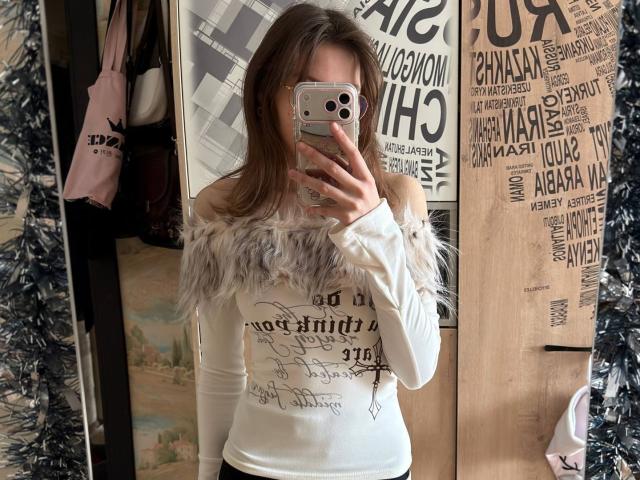 Delfina Auble, de pie frente a un espejo, tomando una selfie con un iPhone. Llevando una blusa blanca con un corte que deja el hombro al descubierto, con letras negras y detalles de piel sintética gris. Pósters y bolsas con diseños atractivos. Increíblemente sexys..