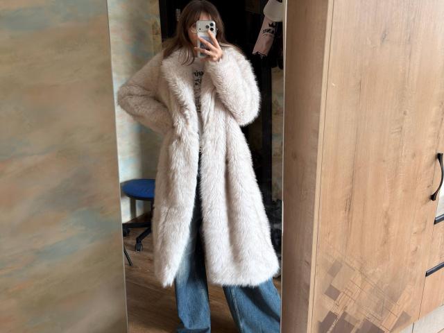 Foto van Delfina Auble, staand voor een spiegel, gekleed in een pluizige witte jas over blauwe jeans, met een telefoon in haar hand. Met één hand, waarbij hun gezicht gedeeltelijk bedekt is. Heel sexy, jas die je penis verbergt..