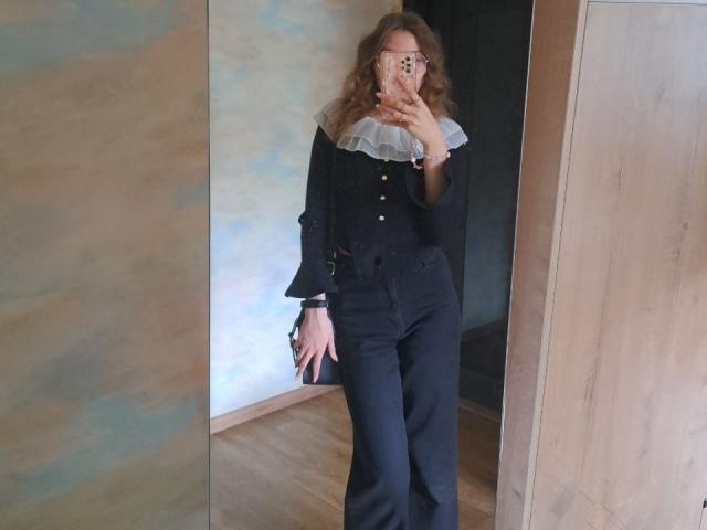 Photograph of DelfinaAuble, wearing a black button-up shirt with a white lace collar, black pants, and glasses, standing in a doorway, holding a phone, with visible sexual tension.