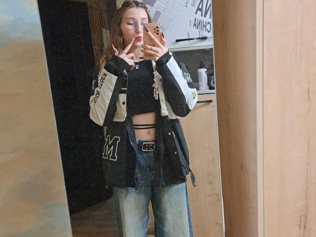 Delfina Auble está em frente a um espelho, tirando uma selfie com o celular. Ela veste uma camiseta curta preta, uma jaqueta esportiva preta e branca e calças jeans azuis. A boca dela está aberta. A mão dela cobre parte do rosto. Os mamilos dela são visíveis através da blusa cropped..
