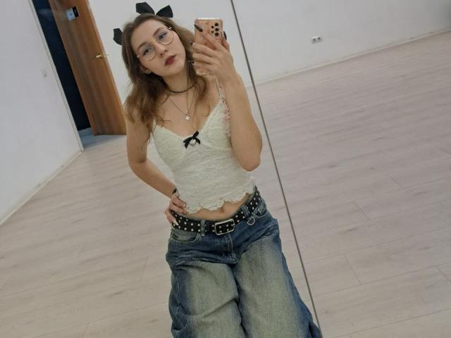 Delfina Auble staat voor een spiegel, met in haar hand een telefoon met een hoesje met kattenoren. Ze draagt een bril, een witte top met kant, blauwe jeans met een riem met studs, en zwarte kattenoren. Haar hand rust op haar heup, waardoor haar buik zichtbaar is. Verdomd sexy..