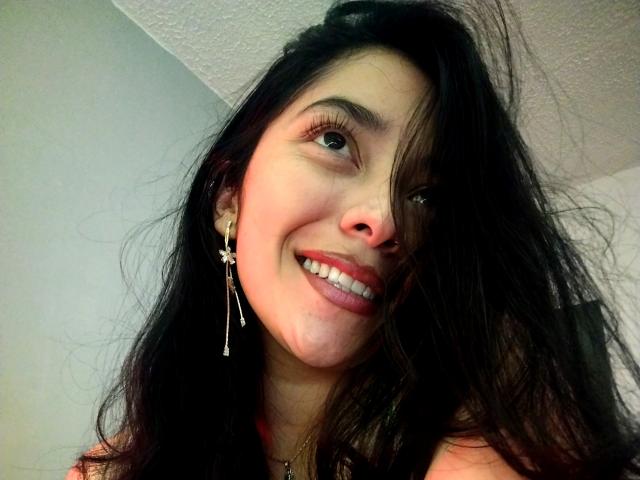 Primo piano di ChantalBernadette, che sorride con i capelli scuri e disordinati che le coprono parzialmente il viso. Ha delle lunghe ciglia, rossetto rosso e indossa degli orecchini a forma di stella. I suoi occhi sono luminosi e leggermente seducenti..