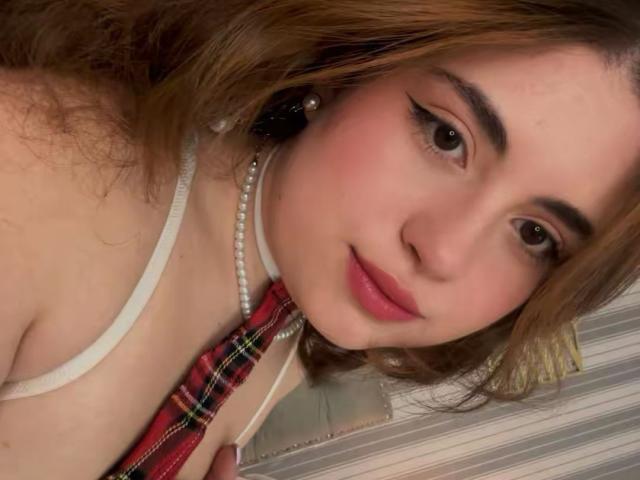 Close-up van VicluX, een jonge vrouw met lichte huid, bruin haar en zwarte eyeliner. Ze draagt een witte band, een parelketting en een rode geruite das, en leunt voorover met een verleidelijke glimlach..