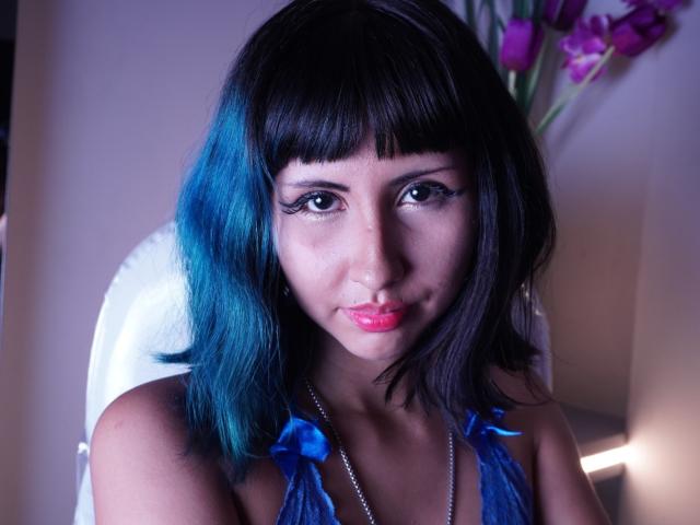 Photographie en gros plan de KleoLennox, une femme aux cheveux bleu foncé, avec une frange, et un rouge à lèvres rouge vif, portant un haut bleu sans bretelles. ornées de rubans bleus, regardant directement l'appareil photo avec une expression séduisante.