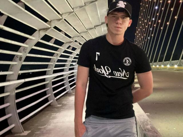 Fotografie von Sebastia Quesada, die nachts unter einem Metallgeländer steht. Er trägt eine schwarze Baseballkappe, ein schwarzes T-Shirt mit weißer Schrift und graue Shorts. Starke sexuelle Anziehung, Penis sichtbar.