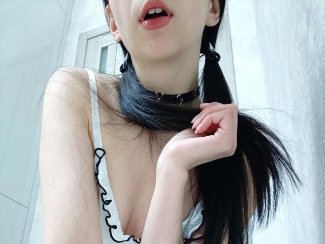 Close-up of WollyMolly with a black choker, white frilled top, and red lips, holding her long dark hair. Her cleavage is visible. Bright white background. Sexual tension, suggestive pose.