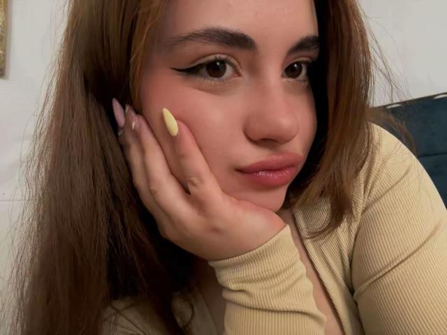 Close-up photo of VicluX, a young woman with long brown hair, resting her hand on her cheek. She has dark eyeliner, yellow nail polish, and a pouty expression. Fucking sexy look.