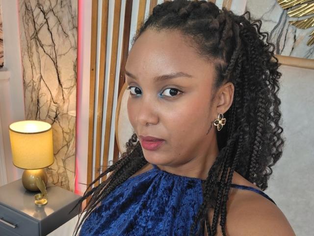 Photograph of TheaBarker with medium brown skin, curly black hair, and red lipstick, wearing a blue lace halter top, looking directly at the camera. Warm light from a lamp in the background.