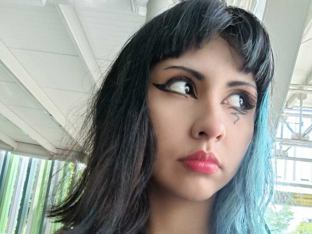 Foto em close-up de KleoLennox com cabelo preto na altura dos ombros, delineador preto marcante e batom vermelho vibrante. Ela olha para o lado com uma expressão sutil e sugestiva. Teto branco, visível do chão..