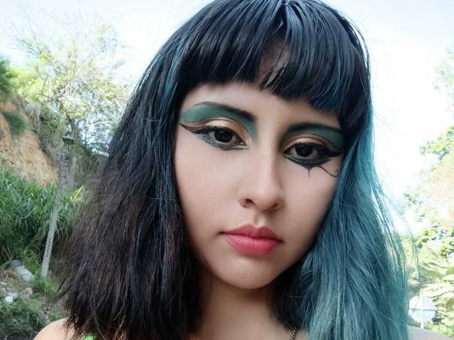 Nahaufnahme eines Fotos von KleoLennox, einer Frau mit schwarzen und blauen Haaren, starkem grünen Lidschatten und roten Lippen. Ihr intensiver Blick hat eine subtile sexuelle Spannung, mit einem natürlichen Hintergrund im Freien..
