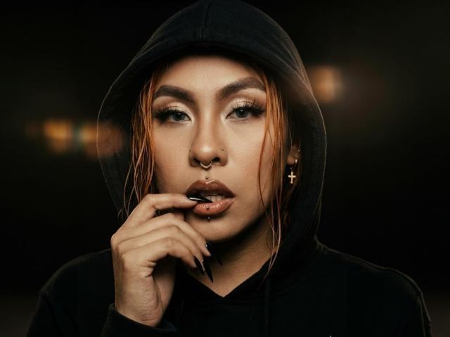 Fotografia ravvicinata di SharomCastle, una donna asiatica attraente con capelli rossi e bagnati, che indossa una felpa nera con cappuccio. Mordendosi seducentemente il dito, occhi scuri, orecchino a croce, rossetto nero, sfondo scuro..