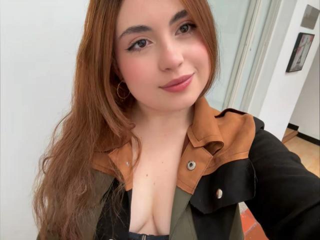 Close-up of VicluX with long brown hair, fair skin, and pink lips, wearing a black jacket with a brown leather collar, exposing cleavage. She smiles slightly, brown eyes looking forward.