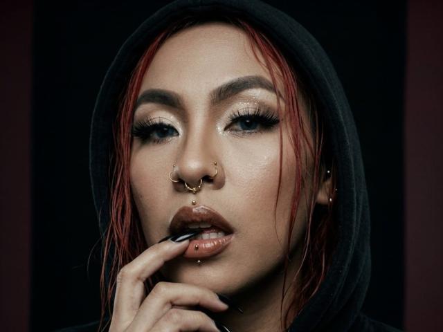 Nahaufnahme von SharomCastle mit rot gefärbten Haaren, Nasenring und starkem Make-up, die verführerisch an ihrem Finger nagt. Dunkel gefärbter Kapuzenpullover, glänzende Haut, intensiver Blick, sexuelle Spannung, leckende Lippen.