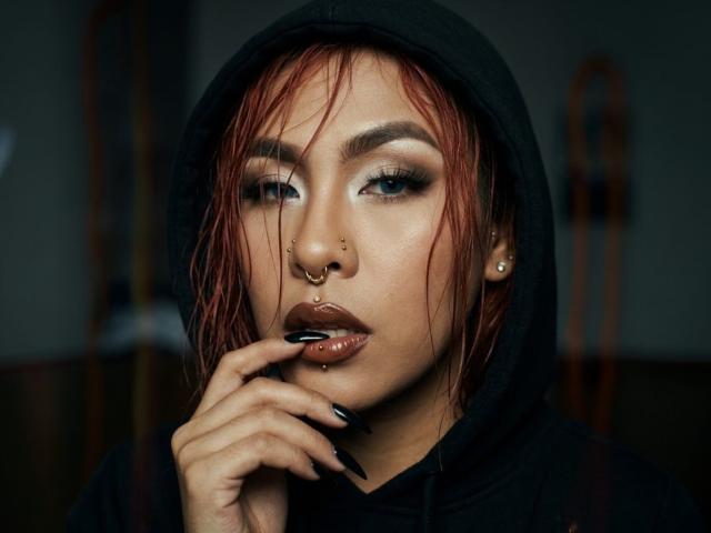 Foto em close-up de SharomCastle, uma mulher atraente com cabelo vermelho molhado, maquiagem pesada e um piercing no nariz, lambendo o dedo. de forma provocadora.
capuz escuro, pele brilhante, olhar intenso e uma expressão sedutora..