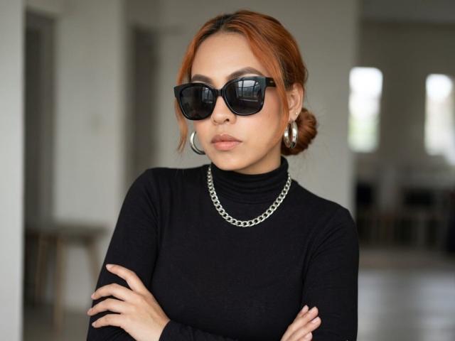 Fotografia de SharomCastle, plano médio, com os braços cruzados, vestindo uma gola alta preta, óculos de sol pretos grandes e uma corrente de prata. colar e brincos de argola. Cabelo ruivo preso em um coque, maquiagem discreta, expressão confiante. Fundo desfocado..