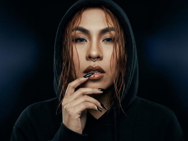 Close-up photo of SharomCastle, wet red hair, black hoodie, nose ring, dark lipstick, black nail polish, hand near mouth, intense gaze, dark background, sexual tension, seductive pose, gothic style.