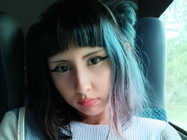 Close-up of KleoLennox, Asian woman with black hair in double buns, heavy black eyeliner, and white ribbed top. She stares directly at the camera with a serious expression. Bright light on the right.