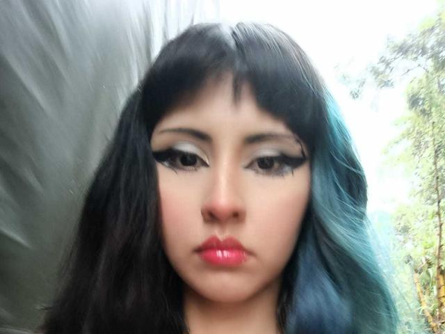 Close-up of KleoLennox, an Asian woman with long black hair, heavy black eyeliner, and bright red lipstick. Blurred background. Her gaze is direct, lips slightly parted, exuding sexual tension.