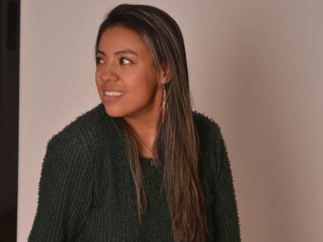 Photograph of MeganFerrerr with dark brown skin, long straight black hair, wearing a black knitted sweater, looking to the side with a smile, showing subtle sexual tension with confident pose.