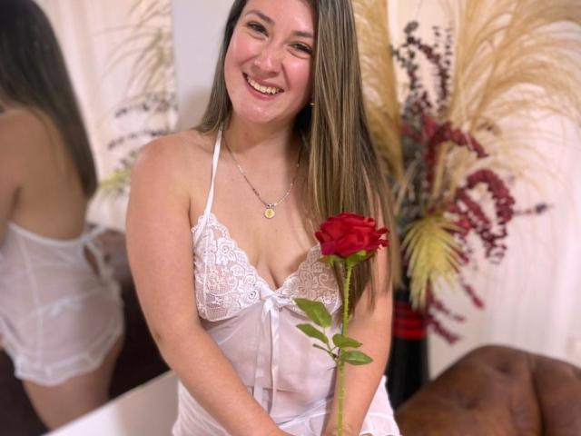 Antonella, con lunghi capelli castani, sorridente, indossa un top di pizzo bianco, tiene in mano una rosa rossa, e porta una collana. ciondolo dorato, riflesso della sua schiena, erba di pampa sullo sfondo.