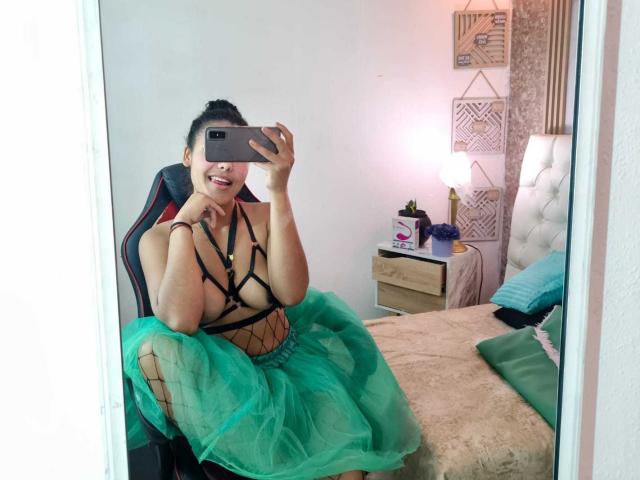 AriaVega, sitting on a bed, taking a mirror selfie. Wearing a black strappy top and a green tulle skirt, fishnet detailing. Smiling, holding phone, visible cock outline. Bedroom setting.