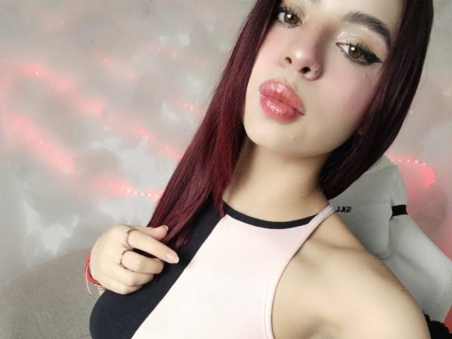 Close-up photo of NiickyySweet, a sexy Latina woman with long dark hair, glossy pink lips, and large brown eyes. She's wearing a black-and-white halter top, touching her hair seductively. Bright pink and white bokeh background.