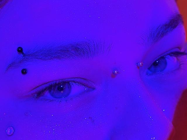 Close-up do rosto de MilaDallila, com os olhos fixos e olhando intensamente para frente. A luz azul ilumina sua pele, realçando as piercings faciais pretas na sobrancelha esquerda e acima da sobrancelha direita. Detalhe em vermelho à direita.
