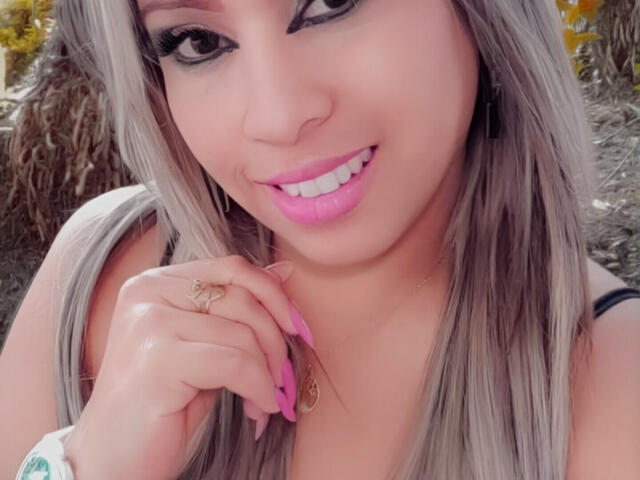 ObedientAndSexy has pink lips, white teeth, long grayish-blonde hair, black eyeliner, pink nails, and a gold ring on her finger, smiling at the camera.