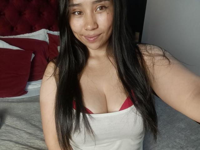 Fotografía de Karollteyes, una mujer asiática con cabello largo y negro, que lleva un top blanco que deja ver las tiras rojas de su sujetador. Sentado en una cama con almohadas rojas. Tiene una leve sonrisa, y sus senos son grandes..