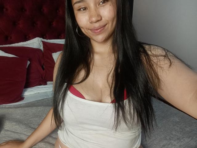 Karollteyes, an Asian woman with long black hair, wearing a white tank top revealing red bra straps, sits on a bed with red pillows and a tufted headboard. She smiles slightly.