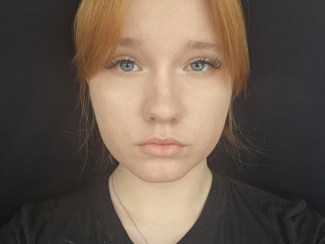 Close-up portrait of GilmaNewhouse with pale skin, light blue eyes, and straight, reddish-orange hair. She has long, dark eyelashes and a neutral expression. She wears a black V-neck shirt. The image focuses on her face, showing slight freckles and a smooth complexion. The background is solid black, making her features stand out. The lighting is even, highlighting her facial details. Her hair is parted in the middle.