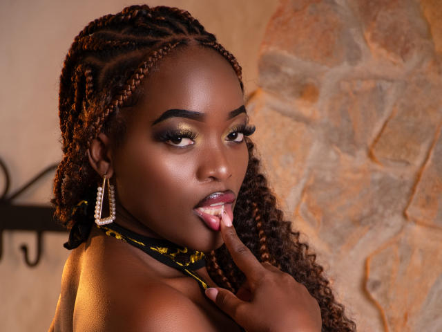 Close-up photo of ZenyaEbony, dark-skinned woman with braided hair, licking her finger seductively. She wears large hoop earrings and a black choker, with warm, textured stone wall background.