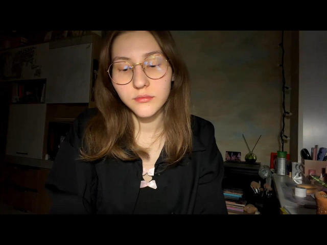 Photograph of DelfinaAuble with light skin, brown hair, and round glasses, wearing a black shirt. She has a serious expression, with dimly lit background featuring a cluttered desk. Fucking sexy.