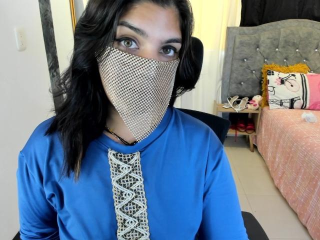 AniisaAli, portant un haut bleu à manches longues avec un col blanc à motifs, a un masque facial en maille métallique qui couvre son nez. et la bouche. Contexte : lit avec une couette rose, une tête de lit grise et un oreiller jaune..