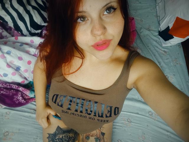 Close-up of SophieHill, pale-skinned woman with red hair, wearing a brown tank top with "OSTRACODE" text, displaying a tattoo on her lower abdomen, looking up with pink lips, slight smirk.