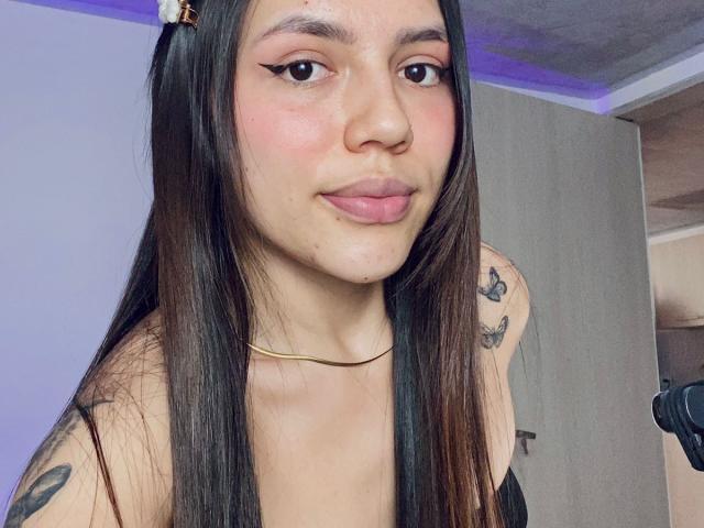 Close-up van SophiaMarx met lang zwart haar, roze oogschaduw en een subtiele glimlach. Ze heeft tatoeages op haar schouders, een dunne gouden ketting, en kijkt recht in de camera. Heel erg aantrekkelijk..