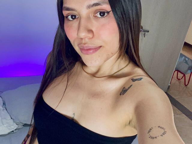 Fotografia di SophiaMarx con lunghi capelli neri, pelle chiara e top nero senza spalline, che mostra tatuaggi sulla spalla e sul braccio. Ha un leggero sorriso, labbra rosate e un décolleté evidente. Incredibilmente sexy..