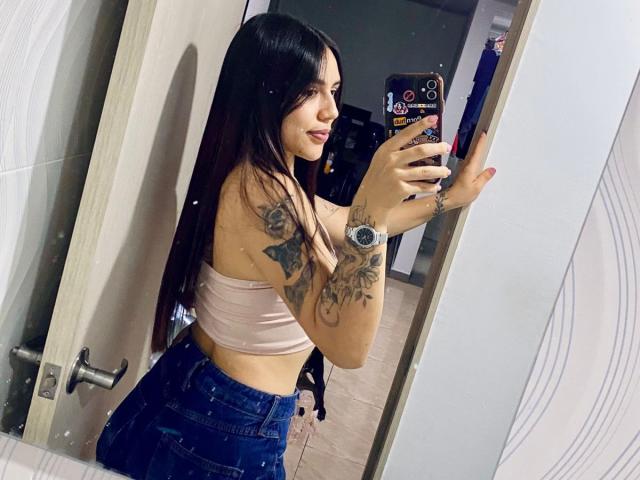 Sophia Marx, mit langen schwarzen Haaren, gebräunter Haut und Tätowierungen, macht ein Selfie im Spiegel. Sie trägt ein pinkes Top und eine Jeans mit hoher Taille, ihre Hand liegt auf ihrem Handy, und sie zeigt ihre Armbanduhr. Verdammt heiß..